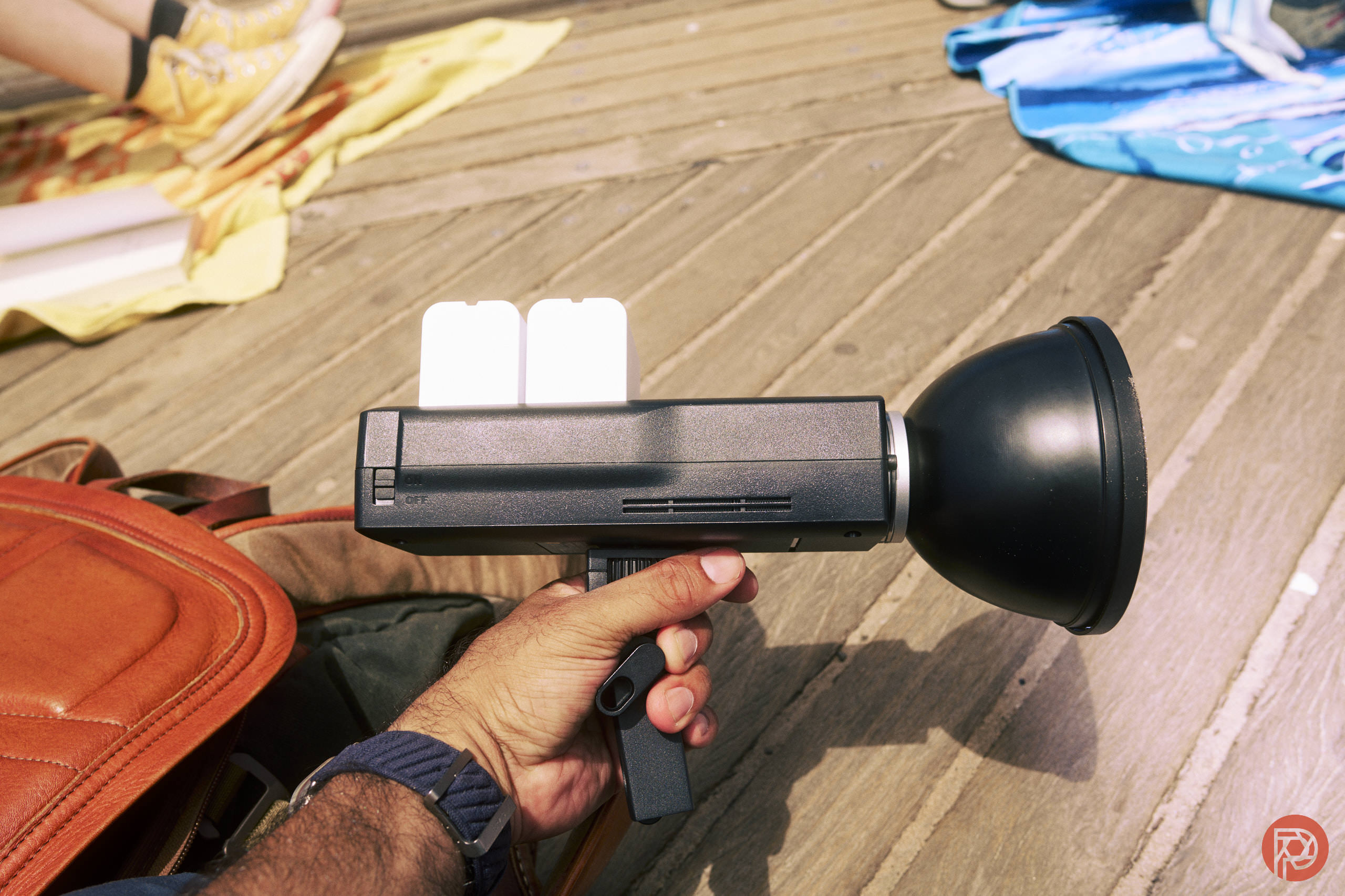 A person holding a handheld black device with a large round front, next to a brown bag on wooden boards.