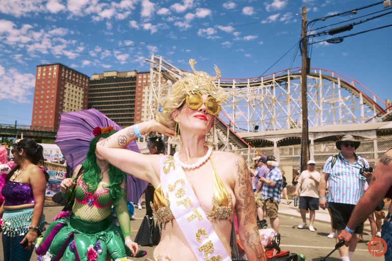Woman in a gold mermaid costume and sash posing at a lively outdoor event near a roller coaster.