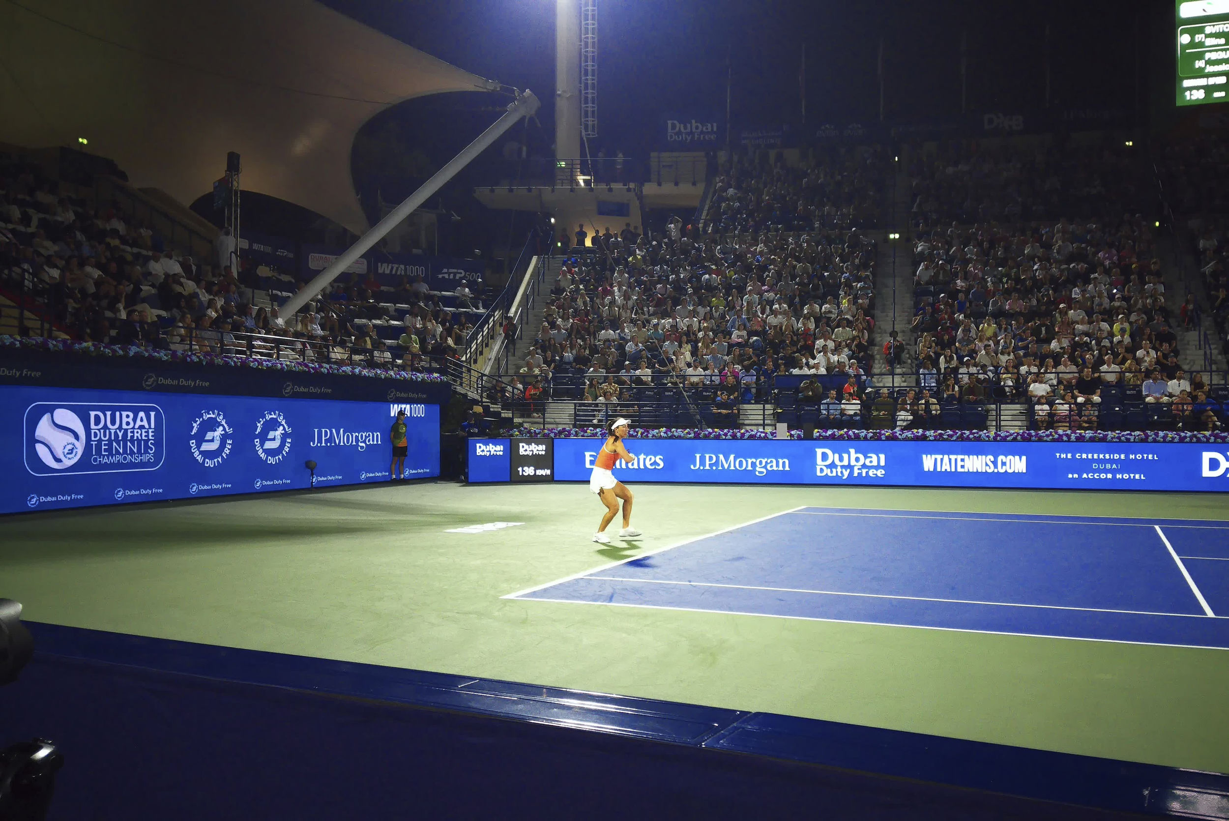 A tennis player serves on a brightly lit court during a night match with a full crowd in the stands.