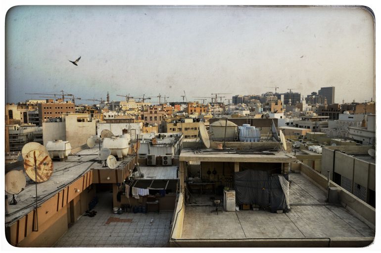 Cityscape with satellite dishes on rooftops, laundry drying, cranes in background, and a bird flying in the sky.