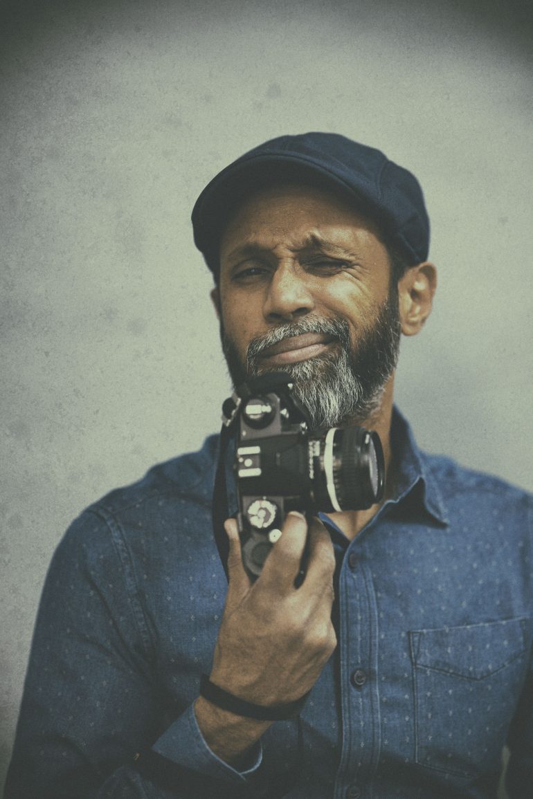 A man in a blue shirt and cap squints while holding a camera up to his face.