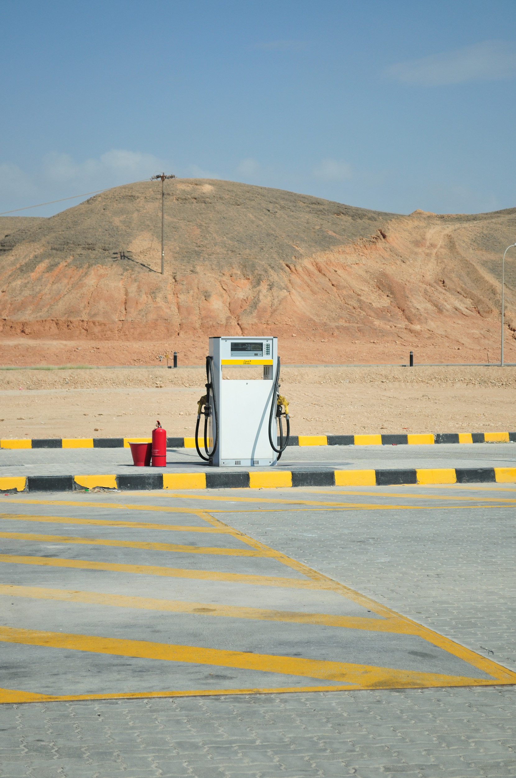 A single gas pump stands on an empty lot with desert hills in the background under a blue sky.