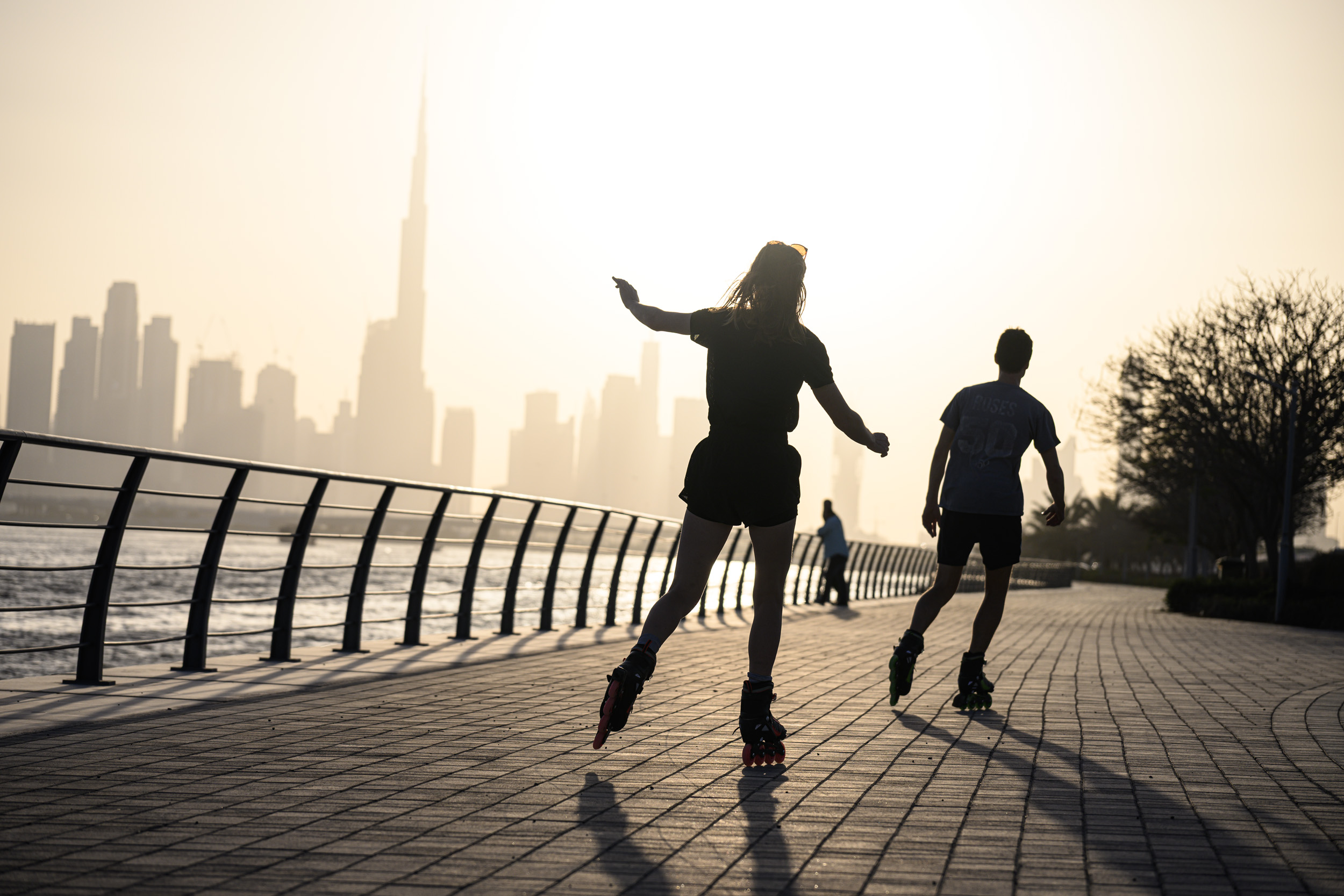 Two people rollerblading along a waterfront path at sunset with a city skyline in the background.