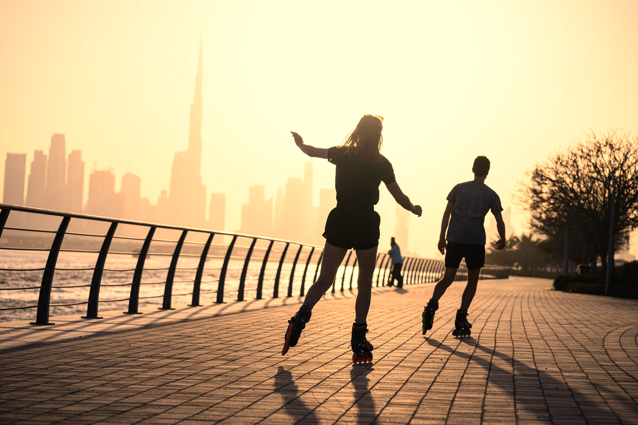 Two people rollerblading on a sunny waterfront path with a city skyline in the background.