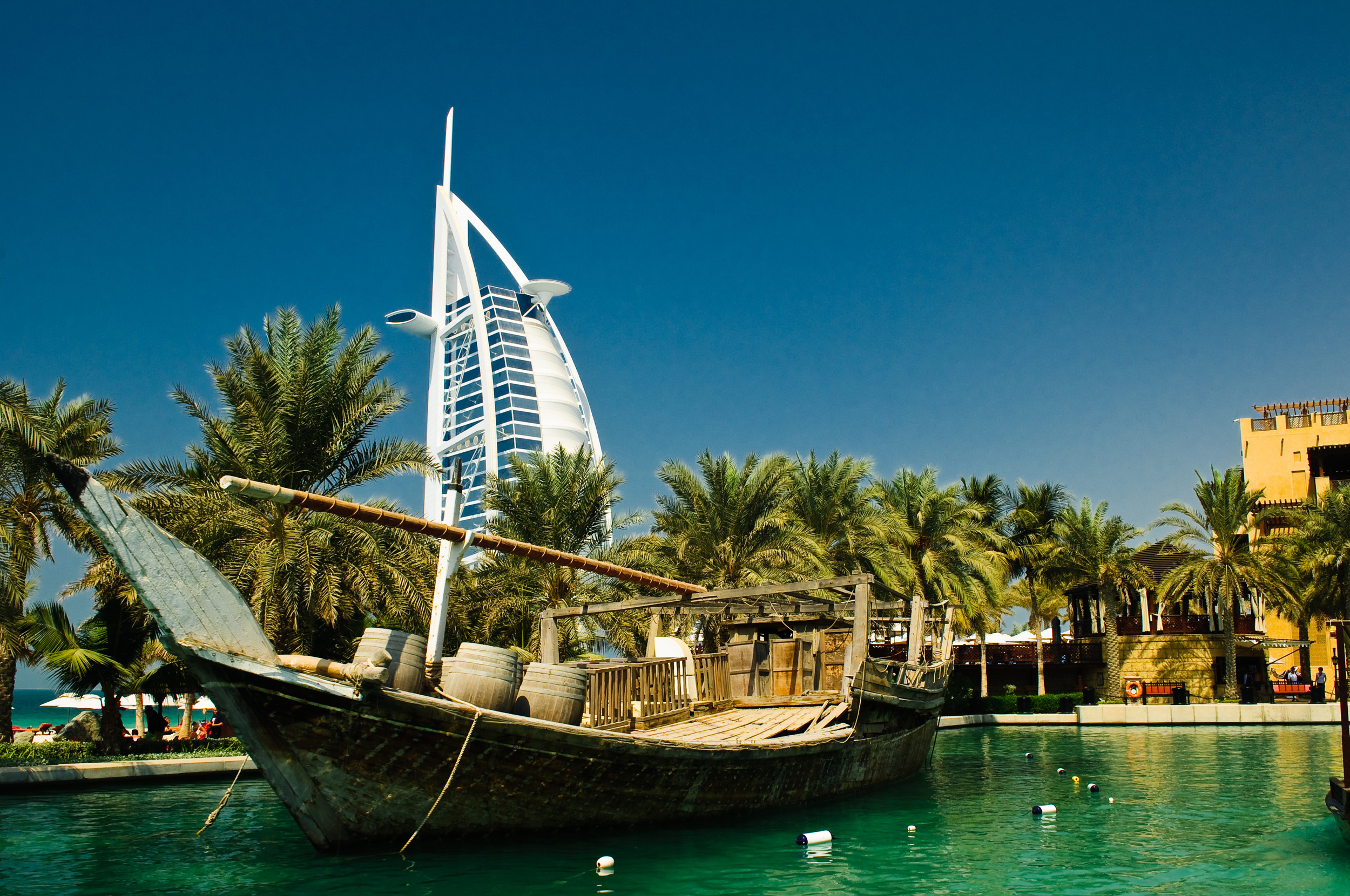 Traditional wooden boat on water with palm trees and Burj Al Arab hotel in the background under a clear blue sky.