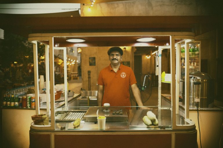 A man stands behind a street food cart with corn and drinks, wearing a red shirt and cap, at night.