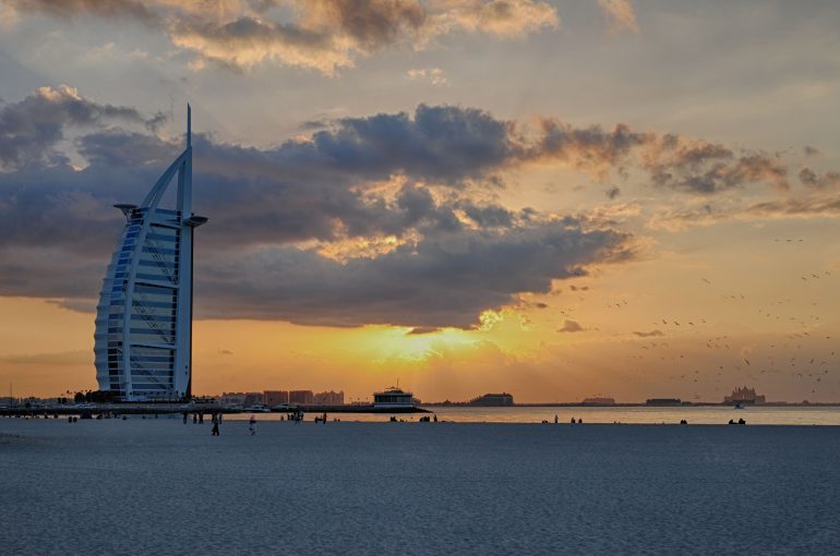 Burj Al Arab at sunset with clouds, sandy beach, and a few people and birds in the distance.