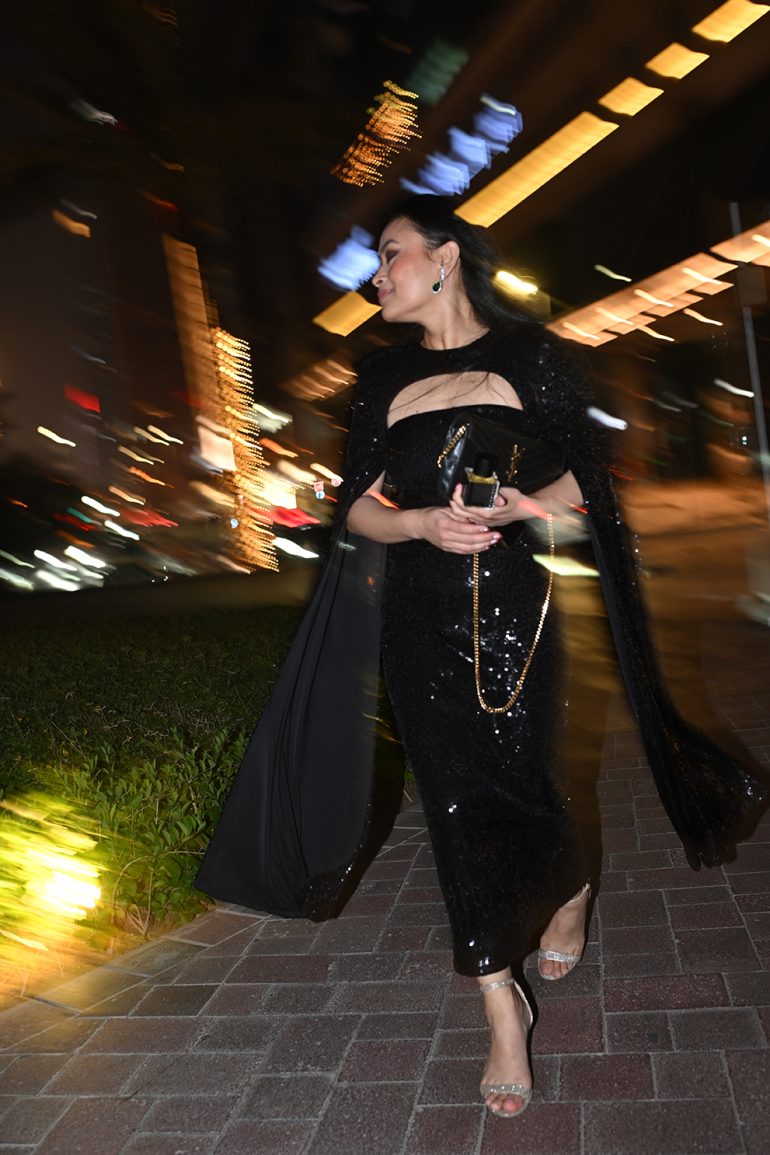 Woman in a sparkling black dress walking outside at night, with city lights blurred in the background.