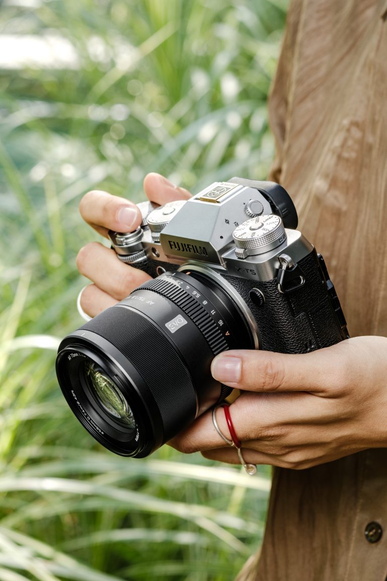 Person holding a Fujifilm camera outdoors with green plants in the background.