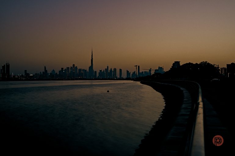 Silhouette of a city skyline at dusk, with calm water and a clear, fading sunset sky.