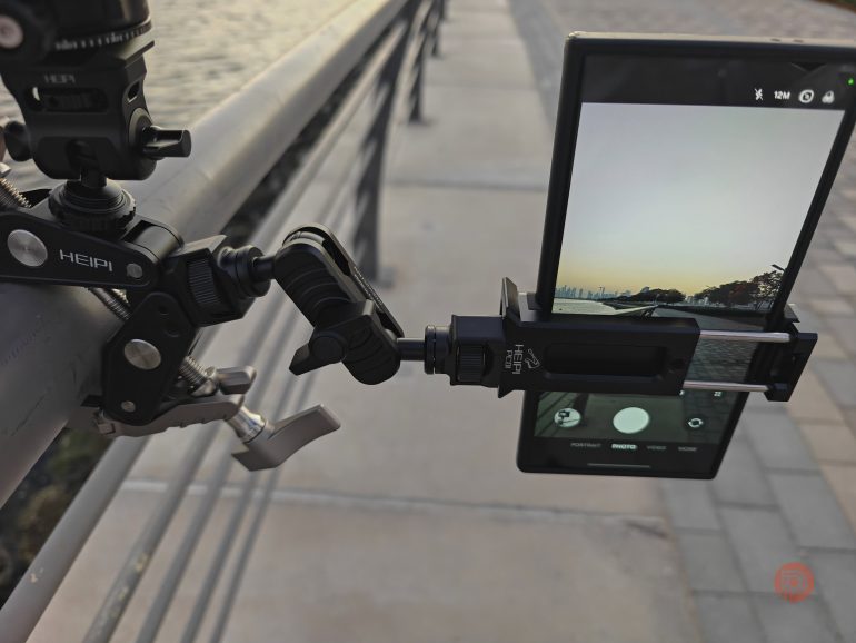 A smartphone mounted on a clamp tripod, capturing a sunset view by a riverside walkway.