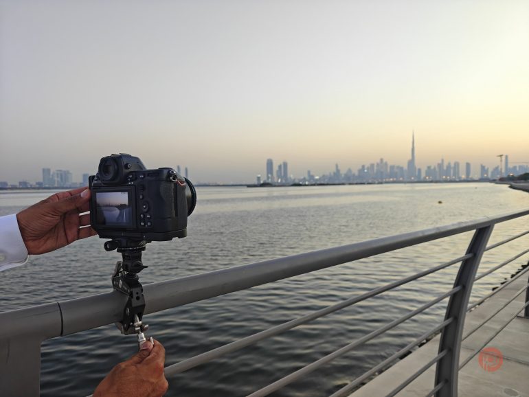 A person secures a camera on a railing, capturing a city skyline across the water at sunset.