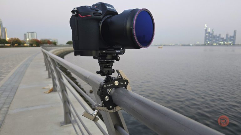 A camera mounted on a rail clamp overlooks a waterfront with city buildings in the distance.