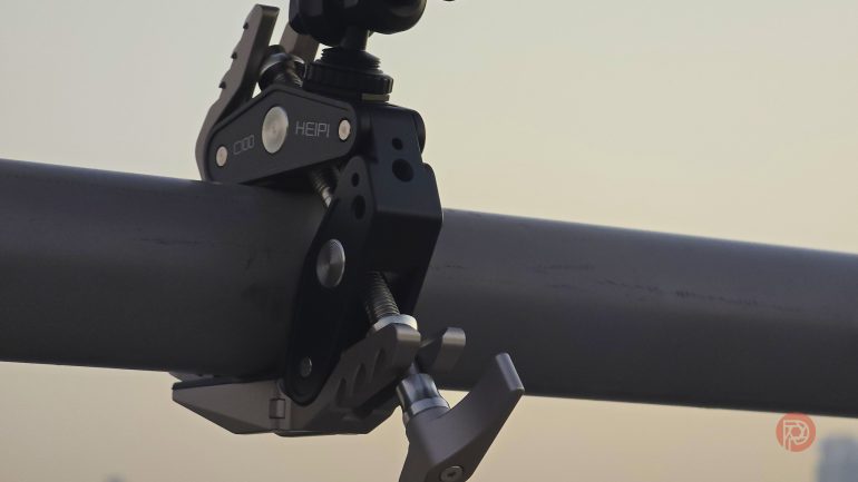 A metal camera clamp attached to a horizontal gray pole against a blurred background.