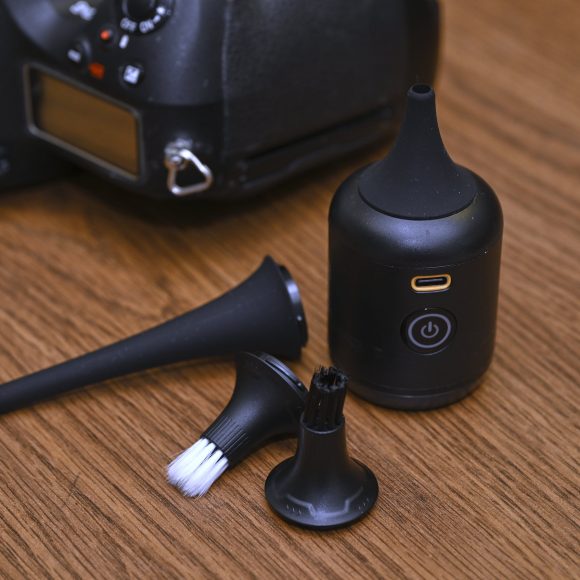 Small black cleaning device with brush attachments, next to a camera, on a wooden surface.