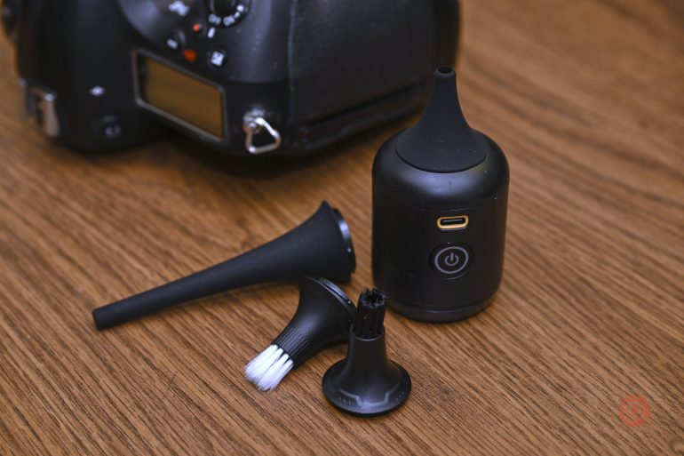 Small black cleaning device with brush attachments, next to a camera, on a wooden surface.