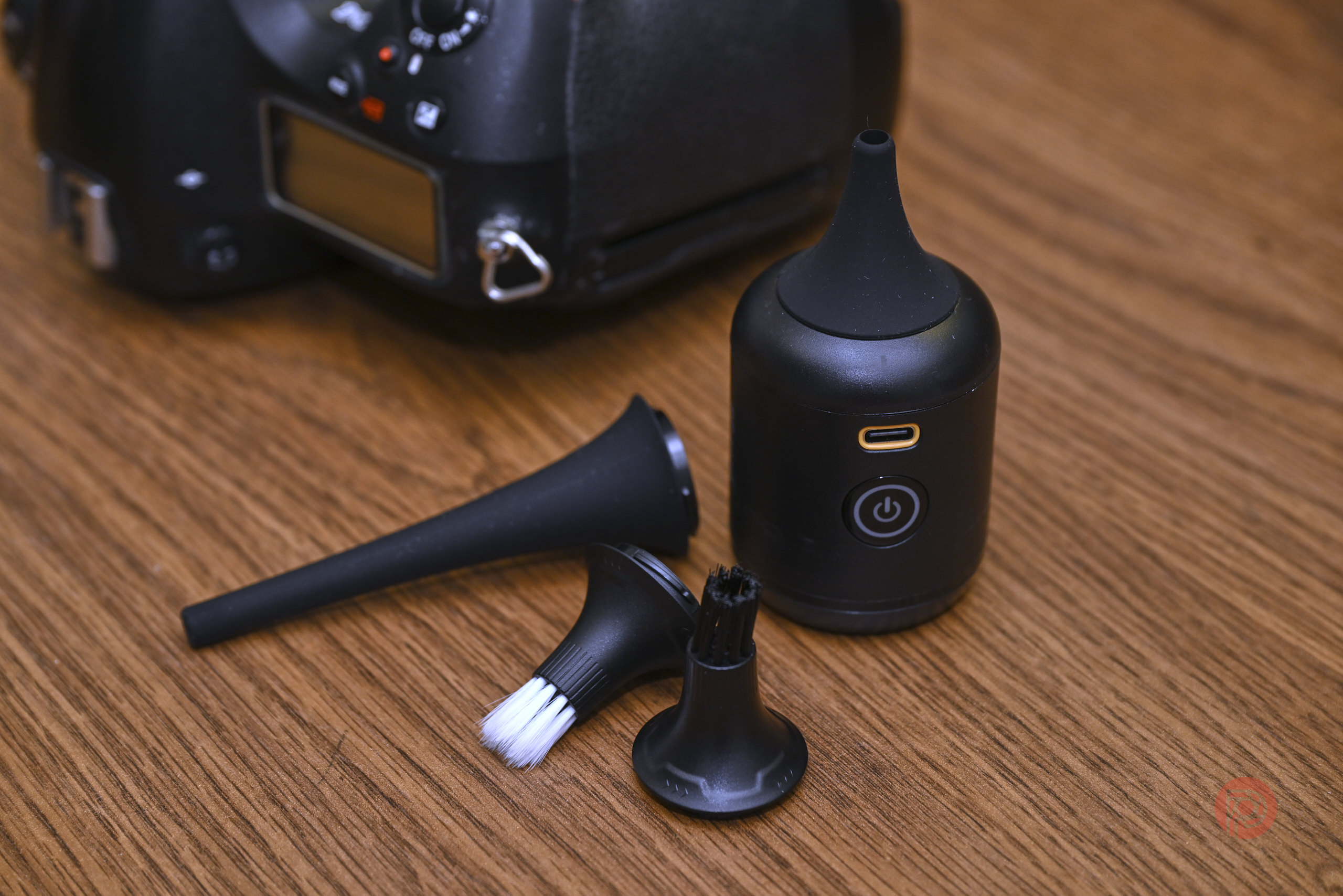 Nitecore BB nano Review: How Did a Blower Get Any Better? Small black cleaning device with brush attachments, next to a camera, on a wooden surface.