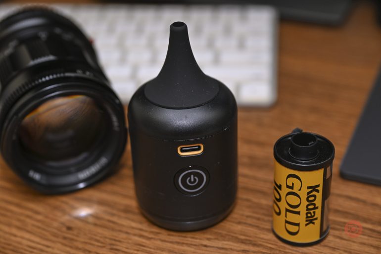 A black device, Kodak film roll, and camera lens sit on a wooden desk near a computer keyboard.