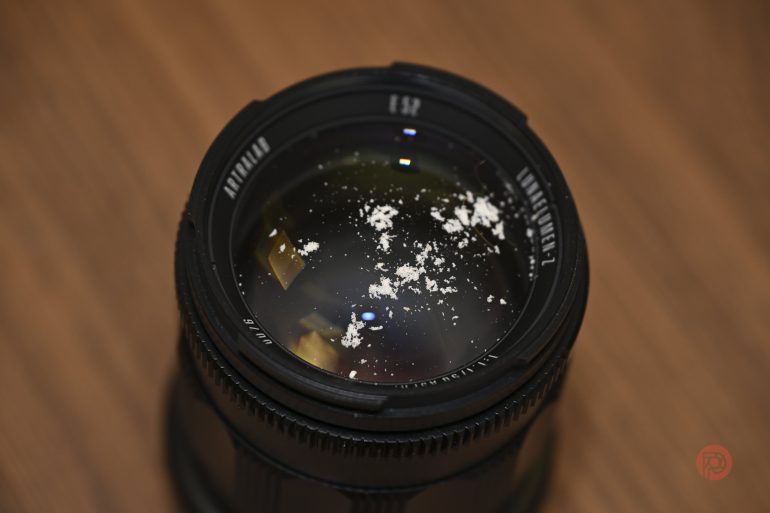 A camera lens with white powdery dust scattered on its glass surface, on a wooden background.