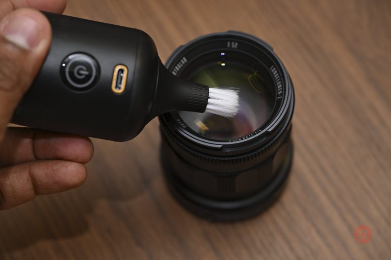 A hand uses a brush tool to clean the lens of a camera on a wooden surface.