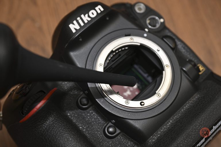 A Nikon camera body being cleaned with a black air blower, lens removed, on a wooden surface.