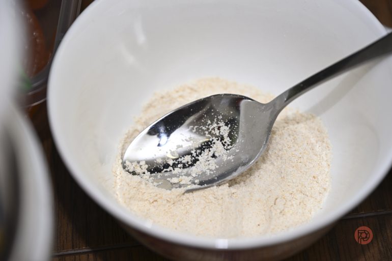 A metal spoon rests in a white bowl containing a small amount of flour or powder.