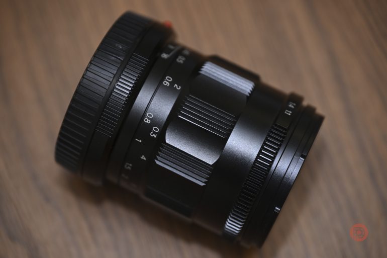 A black camera lens with focus and aperture rings rests on a wooden surface.