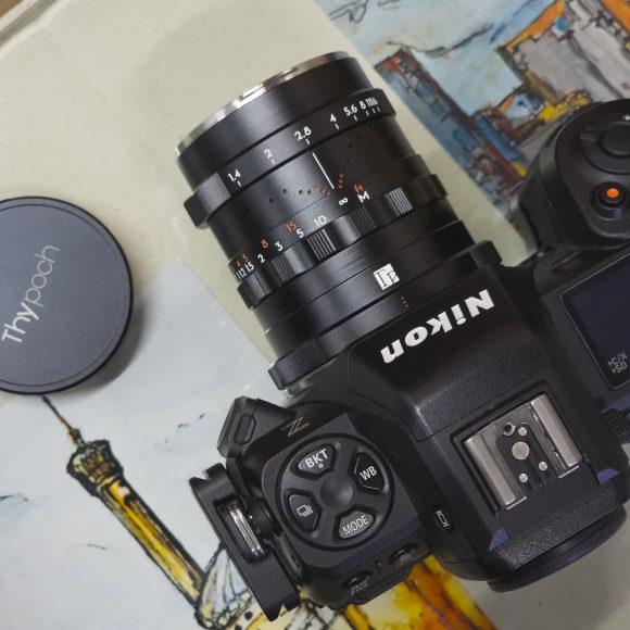 A Nikon camera with a TTArtisan lens and a Thypoch lens cap on a colorful illustrated background.