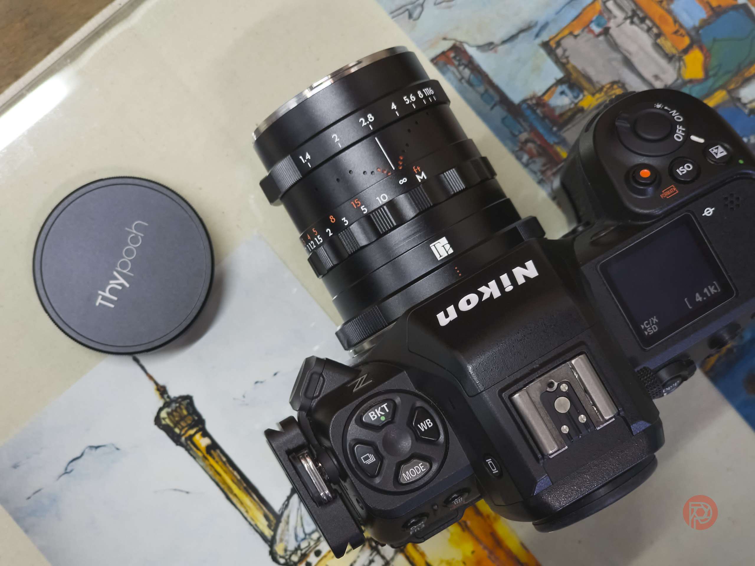 Thypoch Simera 28mm f1.4 Review: The Best Reason To Ditch Your Smartphone Camera A Nikon camera with a TTArtisan lens and a Thypoch lens cap on a colorful illustrated background.