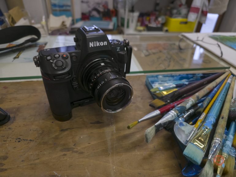 A Nikon camera on a desk beside a scattered set of paintbrushes and art supplies.