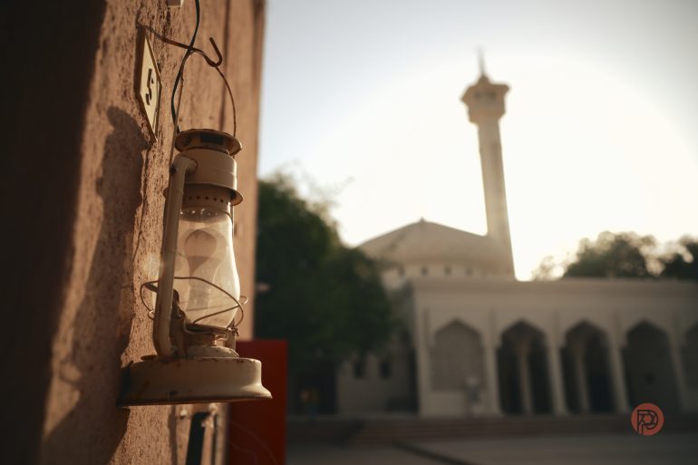 An old lantern hangs on a wall with a mosque and its minaret blurred in the sunny background.