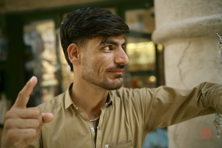 A man in a tan shirt smiles and points upward while standing outdoors near a column.