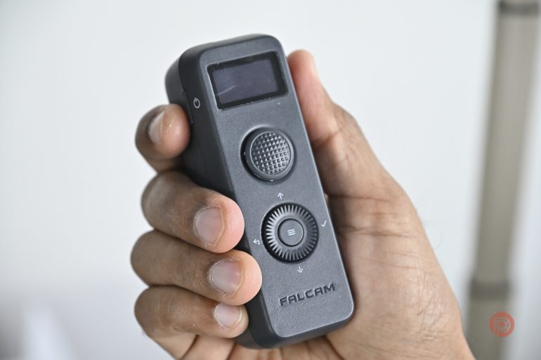 A hand holding a small black FALCAM device with buttons, a screen, and a dial.