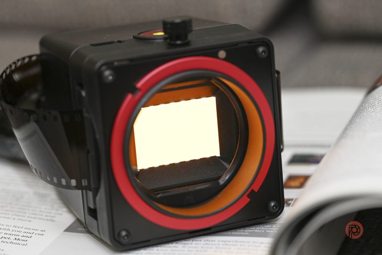 A film negative viewer with a lit screen displays a film strip, resting on open magazines.