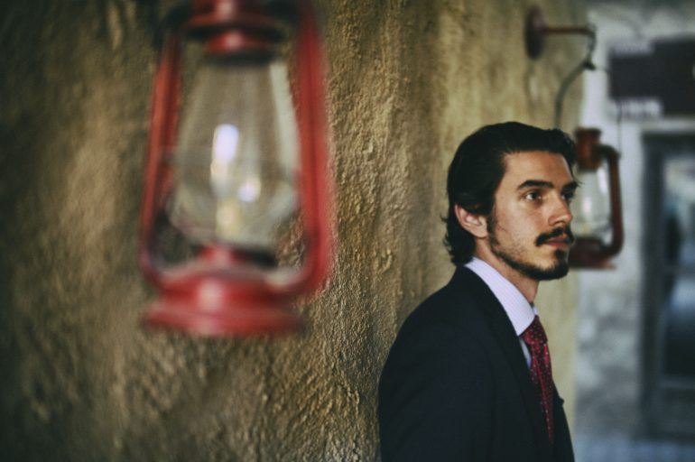 A man in a suit stands by a textured wall with red lanterns, looking thoughtful.