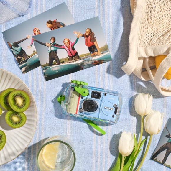 A camera, surf photos, tulips, sliced kiwi, and a drink on a striped cloth with a mesh bag.