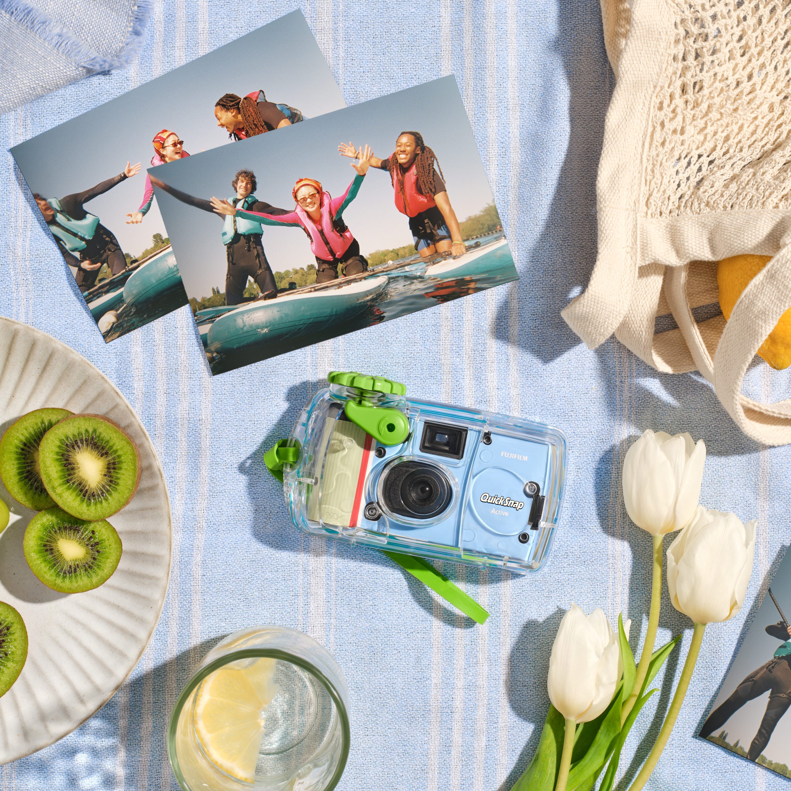 Fujifilm’s QuickSnap Black and White, and the QuickSnap Active Bring Back Nostalgia and Simplicity to Analog Photography A camera, surf photos, tulips, sliced kiwi, and a drink on a striped cloth with a mesh bag.