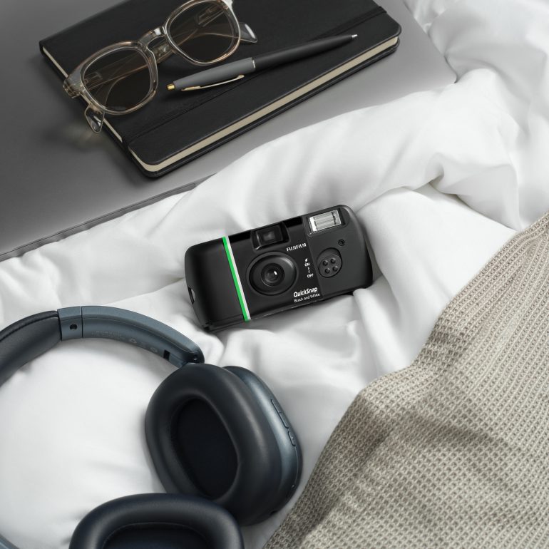 A film camera, headphones, notebook, pen, and glasses on a white bed with a beige blanket.