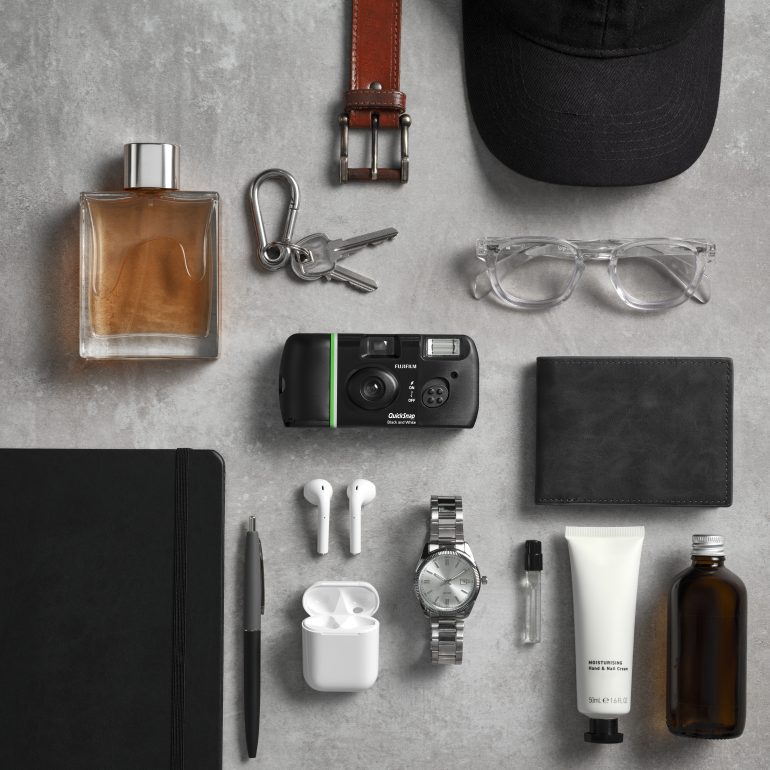 Flat lay of everyday items: camera, watch, glasses, keys, wallet, notebook, pen, earbuds, and accessories.