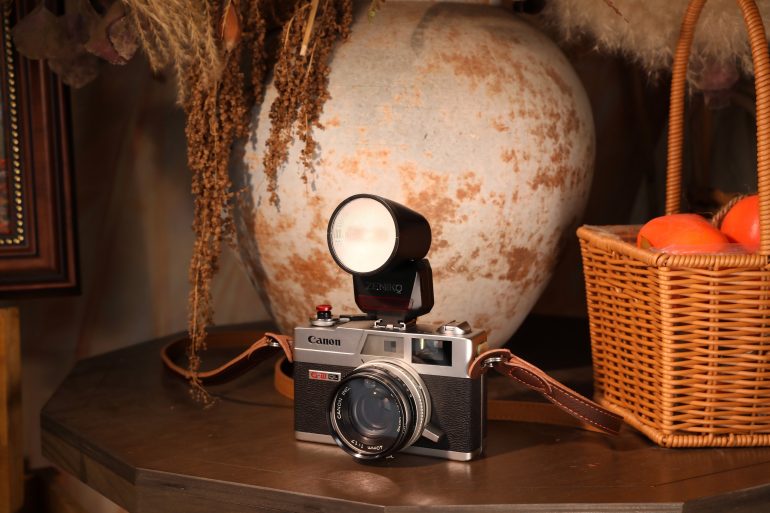 A vintage Canon camera with a flash sits on a table next to a wicker basket of oranges and a large vase.