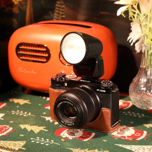 A vintage camera with flash is on a festive tablecloth near an orange radio and a vase with flowers.