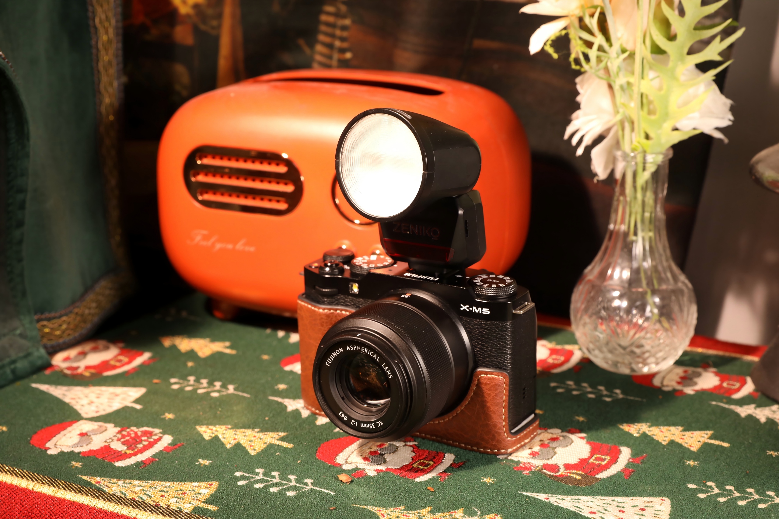 A vintage camera with flash is on a festive tablecloth near an orange radio and a vase with flowers.