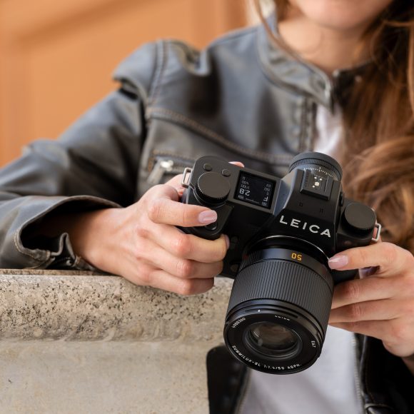 Person holding a Leica camera, wearing a gray leather jacket, standing by a stone ledge.