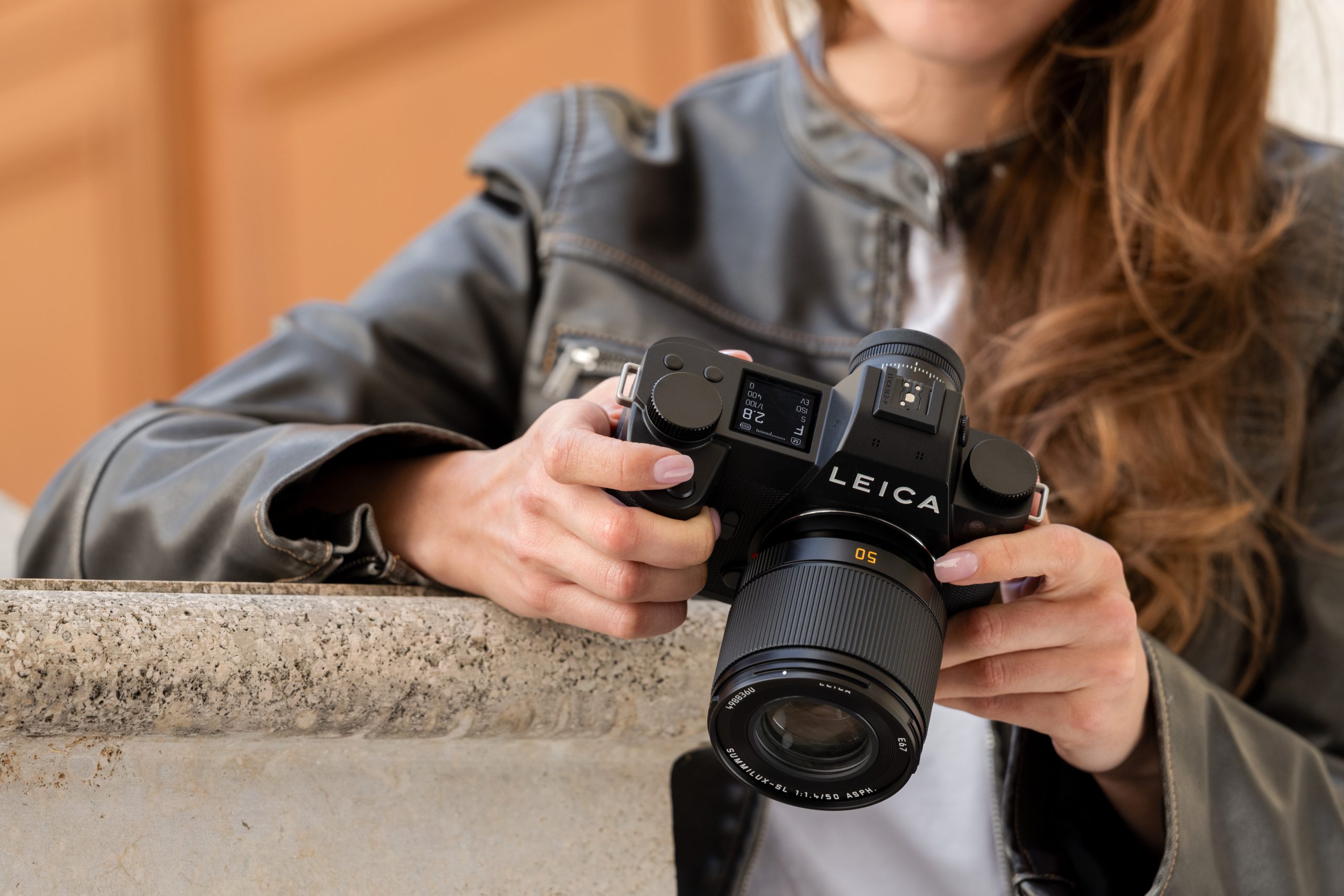 Person holding a Leica camera, wearing a gray leather jacket, standing by a stone ledge.