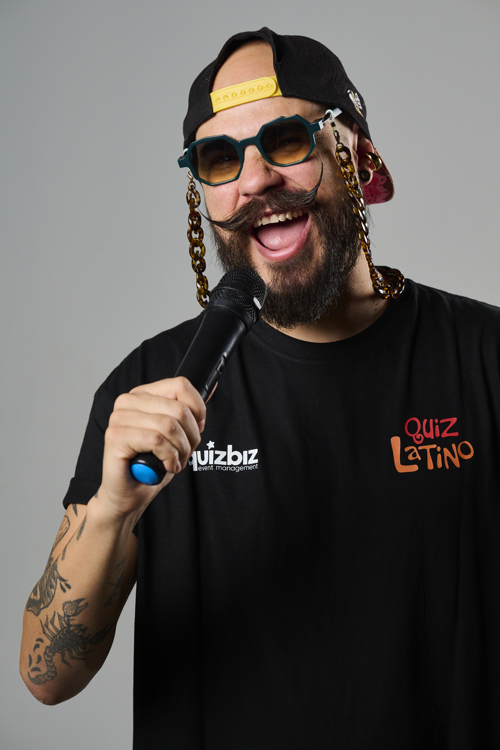 Smiling man in quirky sunglasses, cap, and t-shirt holding a microphone, posing enthusiastically.