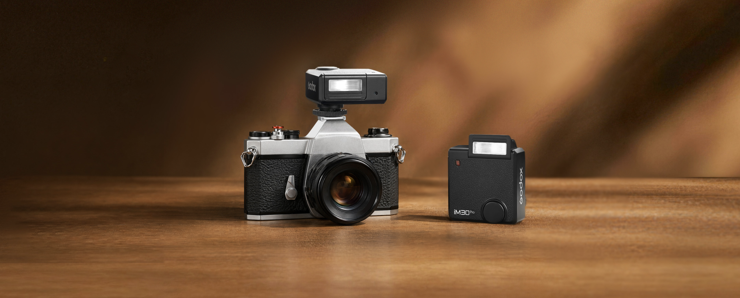 The iM30Pro Is Godox's Latest Pocket Sized Fun Flash A vintage film camera with a flash attached, next to a separate camera flash, on a wooden surface.