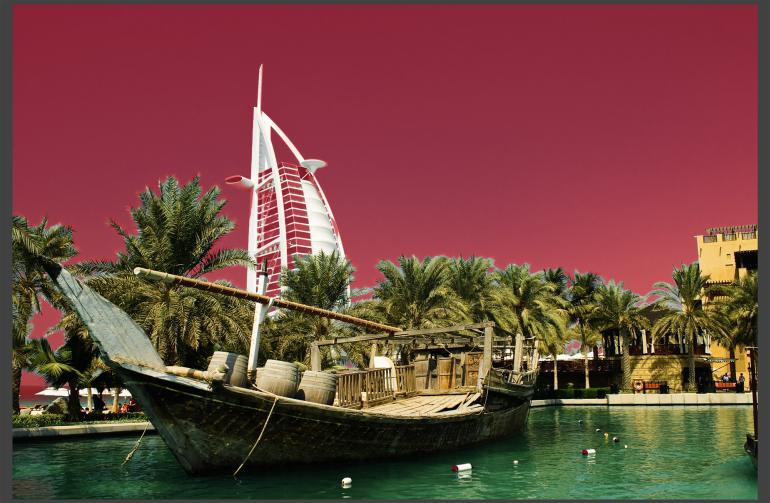 Traditional wooden boat on turquoise water with palm trees and Burj Al Arab against a vibrant pink sky in Dubai.