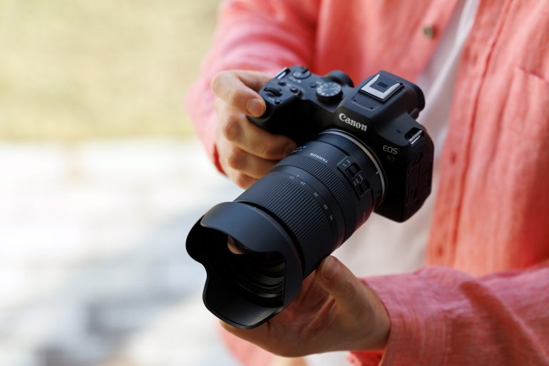 Person in a pink shirt holding a Canon EOS R7 camera with a large zoom lens outdoors.