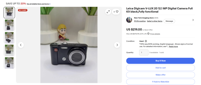 A black Leica digital camera on a white surface, with a small figurine in the background.