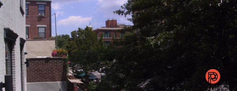 View of city street with trees, brick buildings, and cars on a sunny day; orange logo in bottom right corner.